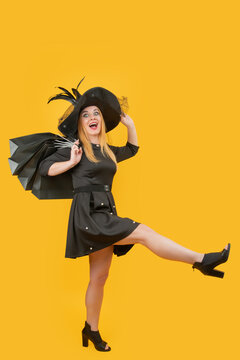 Happy woman holding black shopping bags, wearing a stylish black dress and hat, stepping playfully on a yellow background, expressing excitement and consumerism