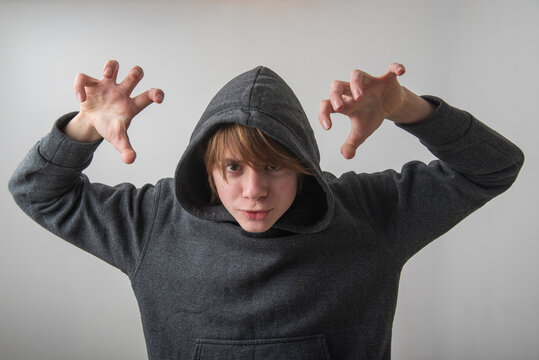 Young boy in gray hoodie with hood up, striking a menacing claw gesture and intense stare, evoking bullying, horror or Halloween costume threat against white studio backdrop