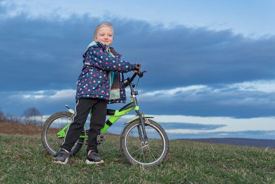 Blond child in a colorful jacket confidently standing with her bicycle on a grassy outdoor hill, expressing happiness and carefree youth exploring nature