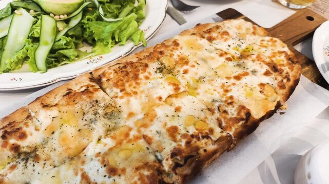 Rectangular cheese pizza bakes with melted mozzarella on long wooden board. Delicious cheese pizza served with herbs offers tasty Italian dinner for friends in local restaurant with drinks.