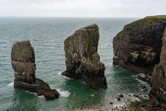 View of the Elegug Stacks, dramatic limestone sea stacks rising from the Atlantic Ocean near rugged cliffs in Pembroke, United Kingdom.