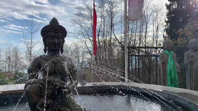 A Buddha statue inside a fountain in a Buddhist temple. Jets of water bathe the statue. In the background are other stone statues, trees, and colorful flags.