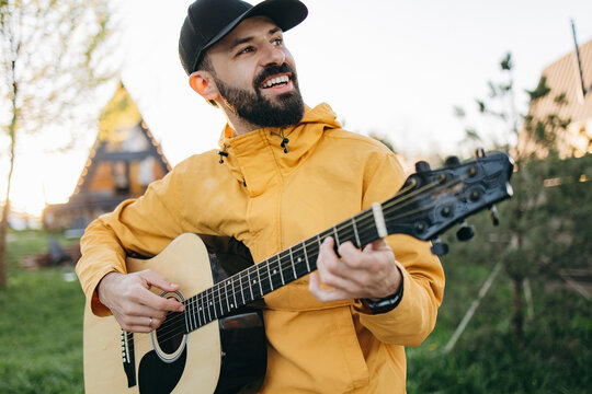 Man with beard plays guitar outside near wooden house smiling warmly at sunset