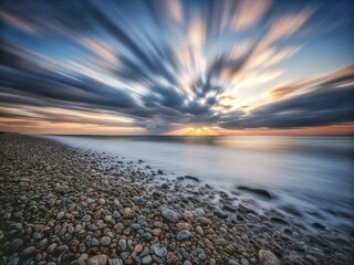 Obraz premium Pebble beach coastline with long exposure smooth water and dramatic sunburst clouds