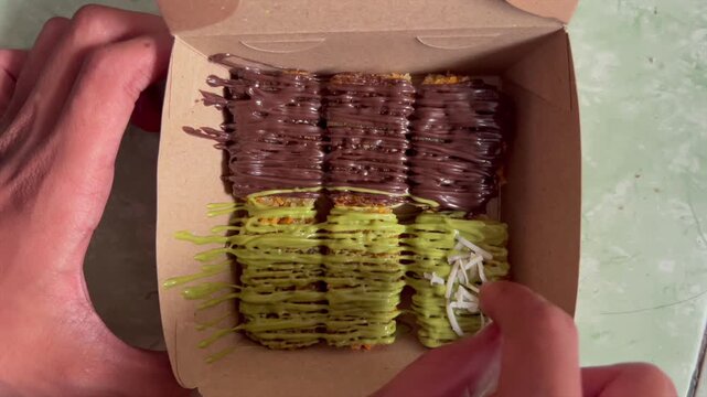 Crispy fried banana nuggets with matcha and chocolate sauce topped with cheese, cookie crumbs, and sliced almonds in a paper box