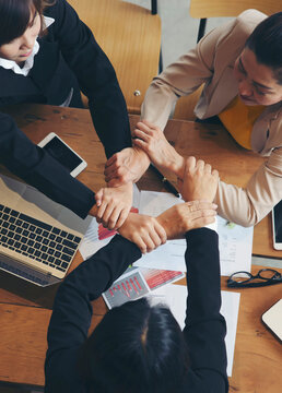 Group of businesswoman joining hands around wooden table during business meeting in indoor office, surrounded by laptops, tablets, documents, and charts, showing teamwork, collaboration, and unity.