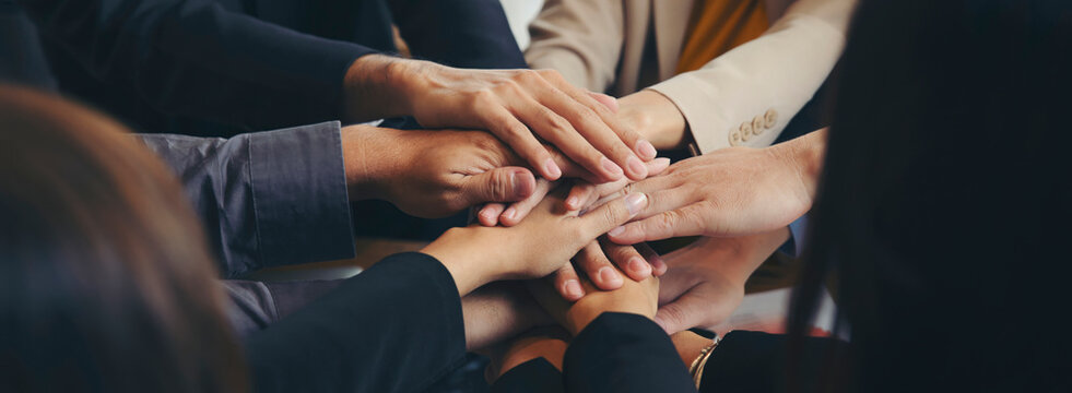 Group of businesspeople joining and holding hands during business meeting at wooden table in office, surrounded by laptops, tablets, documents and charts, showing teamwork, cooperation and unity