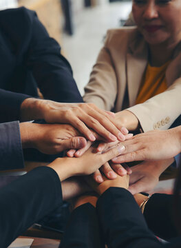 Group of businesspeople joining and holding hand during business meeting at wooden table in office, surrounded by laptops, tablets, documents and charts, showing teamwork, cooperation and unity
