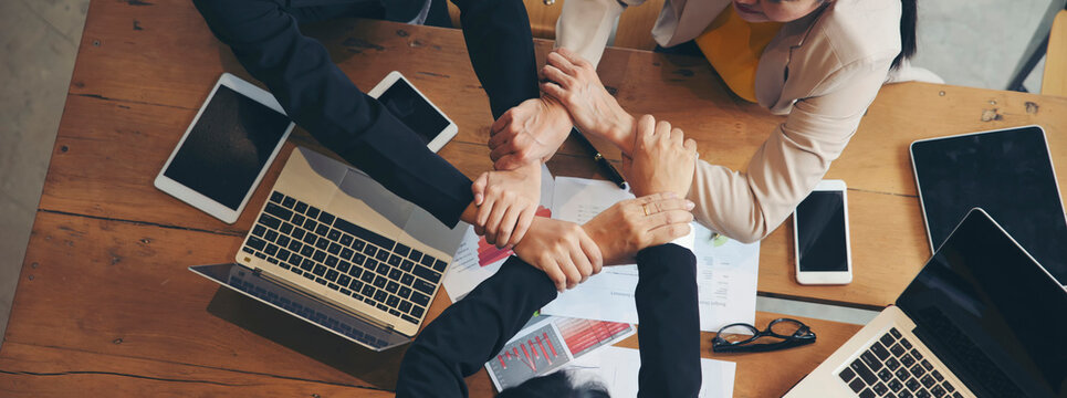 Group of businesswoman joining hands around wooden table during business meeting in indoor office, surrounded by laptops, tablets, documents, and charts, showing teamwork, collaboration, and unity.