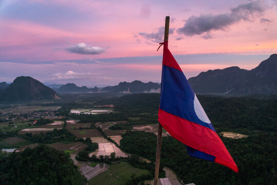 Aerial view of the Laos flag waving on a pole overlooking the lush landscape of rice paddies and karst mountains during a vibrant sunset in Vang Vieng, Vientiane Province, Laos.
