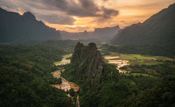 Aerial view of the dramatic limestone karst landscape with lush green rice paddies and a winding path under a golden sunset sky in Vang Vieng, Vientiane Province, Laos.