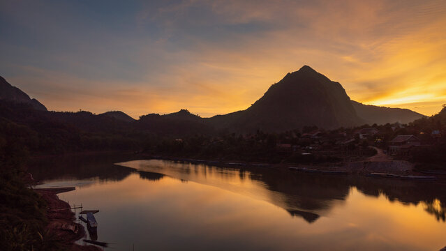 Aerial view of the Nam Ou River flowing past village houses at sunset with mountain silhouettes and golden light reflecting on the water in Nong Khiaw, Luang Prabang Province, Laos.