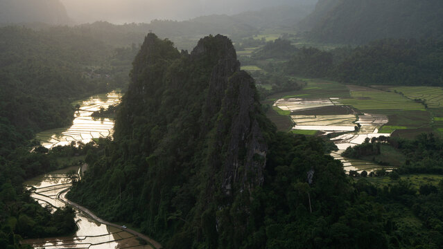 Aerial view of the dramatic karst limestone mountains and lush green rice paddies reflecting the golden sunset light in Vang Vieng, Vientiane Province, Laos.