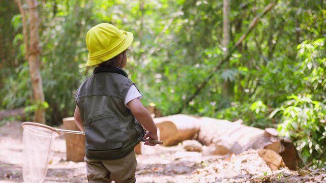A curious toddler wearing an explorer vest and hat holds a butterfly net while on a nature walk, engaging in experiential learning and discovery in the woods.