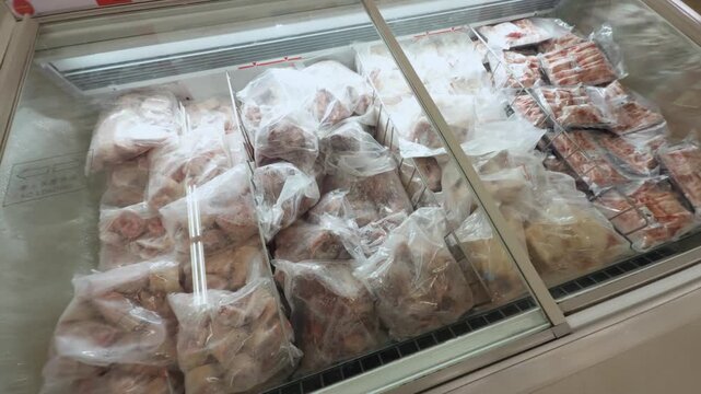 Various cuts of frozen raw meat and poultry are packaged in clear plastic bags, neatly organized within a large display freezer at a grocery store, representing food storage and consumer convenience