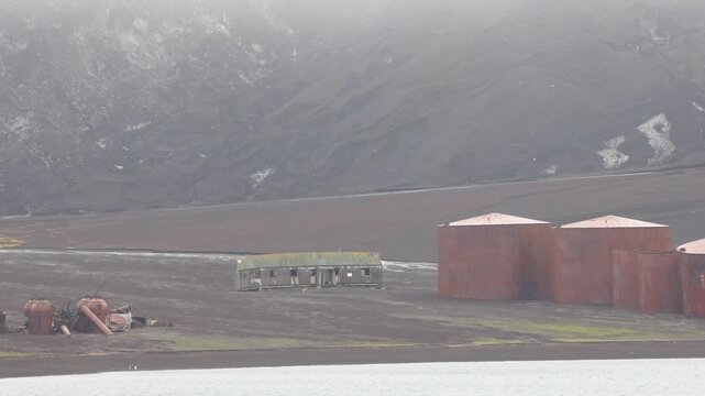 Wide shot of a deserted Antarctic outpost with decaying buildings, rusted storage tanks, and volcanic slopes shrouded in misty, soft natural light. Stark, uninhabited, moody scene.