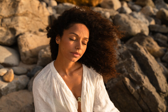 A woman with her eyes closed next to the rocks on the coast at sunset