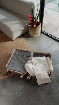 Open pink suitcase packed with white clothes on modern interior floor with plant and sofa nearby
