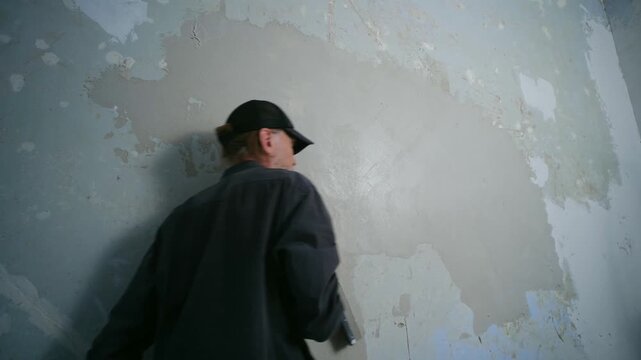 Construction worker in cap and gloves skillfully applying finishing putty on wall with long trowel, working on home renovation project, leveling surface for smooth finish. Plasterer applying plaster