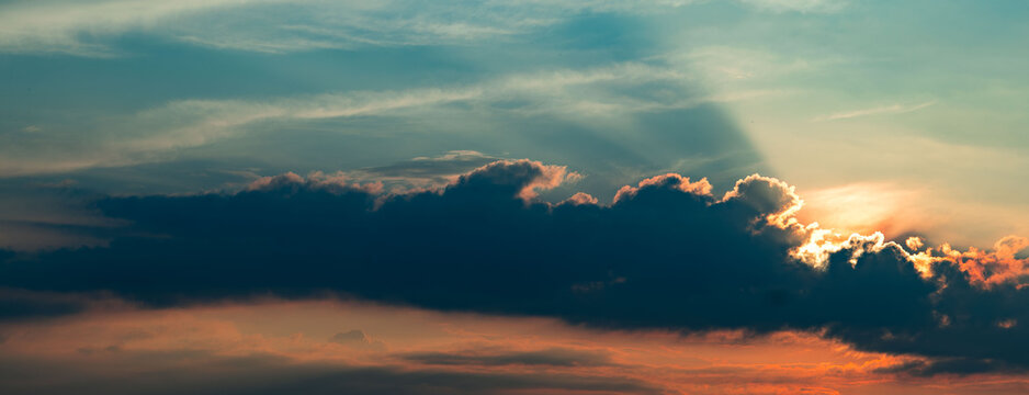 Vibrant evening sky featuring dark, thick clouds illuminated by the setting sun. Golden sunlight breaks through cloud edges, casting radiant beams across a tranquil sky that shifts from warm orange ne
