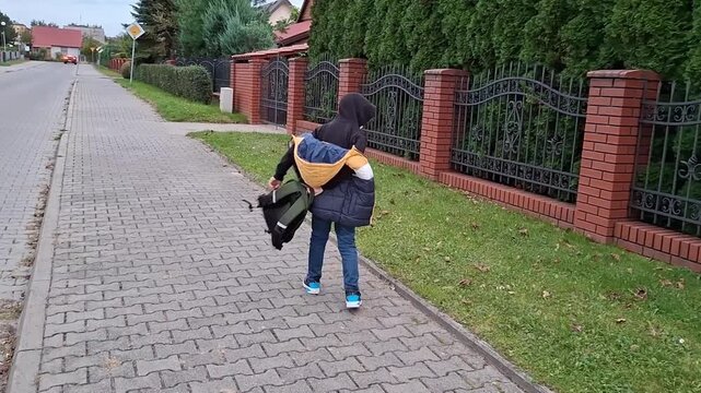 7 year old boy runs down the sidewalk with a jacket slung over his shoulder in a quiet neighborhood