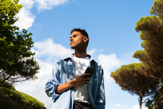 Young Man Standing Outdoors Under Blue Sky With Phone in Hand