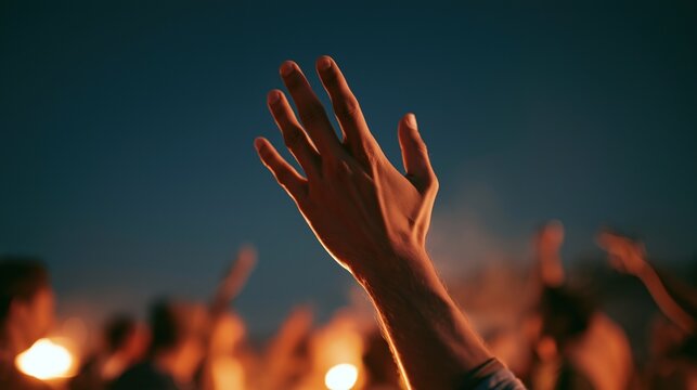Close up of raised open hand reaching upwards lit by warm golden fire light at night outdoor gathering with blurred crowd and glowing bonfire in dark background