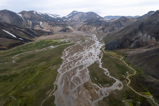 Aerial view of Landmannalaugar featuring a braided river system winding through lush green valleys and rugged volcanic mountains with patches of snow in Rangarthing ytra, Iceland.