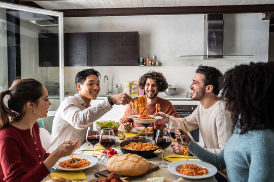 Multiethnic millennial and gen z friends share spaghetti during a lively dinner in a modern home kitchen. Warm group scene celebrates home cooking, hospitality, friendship, and togetherness.