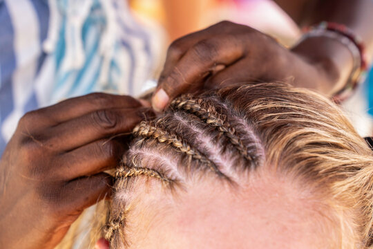 Close-up of a beach hairstylist creating tight cornrow braids on blonde hair during an outdoor summer beauty service. Precise scalp sectioning shows the braiding process for a vacation look.