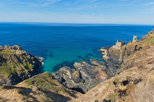 A dramatic coastal landscape shows a deep blue ocean meeting a turquoise cove. Rugged cliffs feature historic stone ruins under a clear sky - Botallack Mine & Engine House, Cornwall, UK