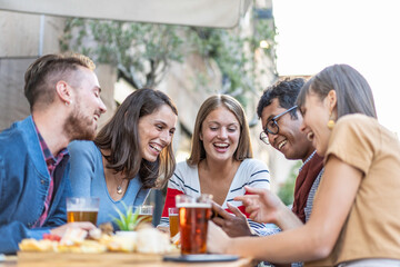 Obraz premium Friends laughing over a smartphone at an outdoor cafe table. Multiethnic millennials and gen z share beer, snacks and funny content on a sunny terrace. Social media concept