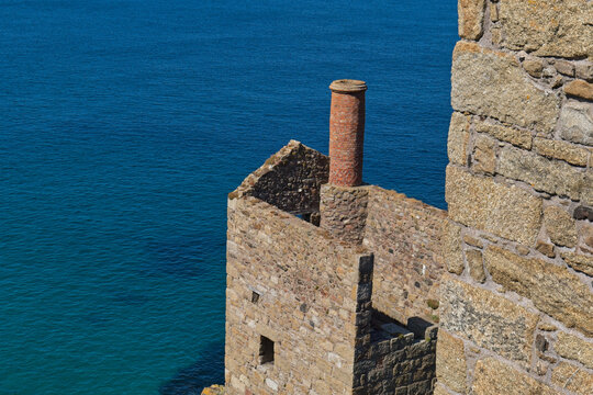 A weathered stone building with a tall brick chimney stands beside a vibrant blue ocean. Part of a thick stone wall is on the right  - Botallack Mine & Engine House, Cornwall, UK