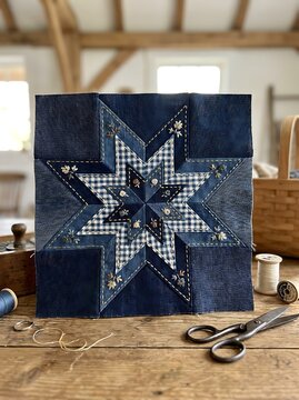 Handmade patchwork star quilt block made from blue denim and gingham fabric with decorative embroidery stitching on a rustic wooden table with sewing tools
