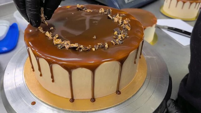 Confectioner decorating caramel drip cake with chocolate crumbs in bakery kitchen. Pastry chef in black gloves finishing glossy mirror glaze dessert on turntable