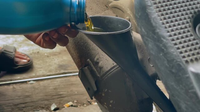Mechanic pouring engine oil from blue bottle through plastic funnel into vehicle component in workshop