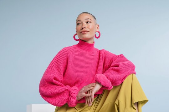 Woman with short cropped hair wearing a pink ribbed knit sweater, sitting poised on a white plinth with copy space for fashion and confidence campaigns