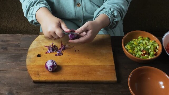A woman's hands peel a red onion with a knife on a cutting board