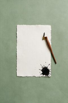 Minimalist flat lay of an oblique calligraphy dip pen and a black ink blot mistake on a deckle-edge paper sheet. Calligraphy writing error concept.