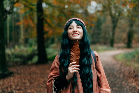 Young smiling woman in warm Shawl Poncho holding fallen leaf and enjoying the moment of calm and freedom in autumn forest walk. Feeling harmony, reunion with nature. Relaxing, mental recovery.