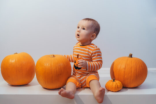 Halloween background with little surprised Caucasian baby in orange costume with lantern jack sitting among pumpkins on white background. Autumn fall harvest festival concept. Copy space
