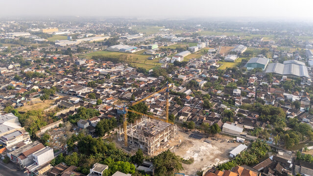 Aerial view of a construction site with a tower crane rising above a dense residential neighborhood with houses, trees, and industrial buildings in Central Java, Indonesia.