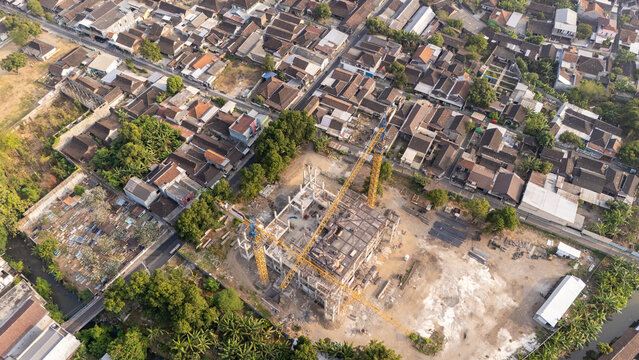 Aerial view of a construction site with a yellow tower crane and concrete framework, surrounded by dense residential rooftops, trees, and a canal in Central Java, Indonesia.