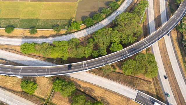 Aerial view of a highway interchange with curved roads, lush green trees, and surrounding agricultural rice fields in Central Java, Indonesia.