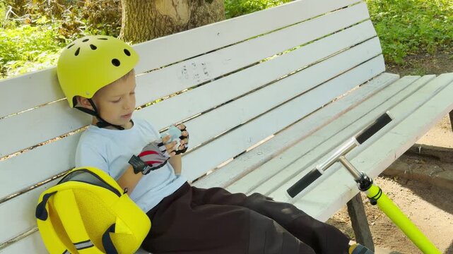 A young boy wearing a yellow helmet rests on a white park bench. He feels calm and relaxed while holding a small gadget in nature.