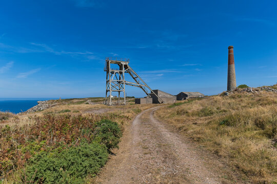 An old mining headframe and chimney stand on a rugged coast under a clear blue sky. A dirt path leads toward the industrial structures on the grassy cliff - Allen's Shaft, Cornwall, UK