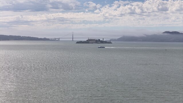 Aerial view of Alcatraz Island and the Golden Gate Bridge in the distance with a ferry crossing the bay under a cloudy sky in San Francisco, California, United States.
