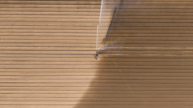 Aerial view of a travelling sprinkler system irrigating crops, with a fine mist of water creating a faint rainbow effect over the dry soil in Mediglia, Lombardy, Italy.