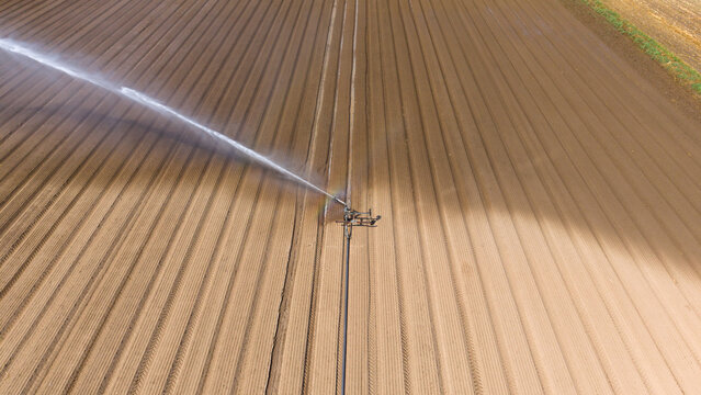 Aerial view of a travelling sprinkler system irrigating brown agricultural fields with furrows in Mediglia, Lombardy, Italy.