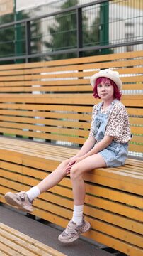 Child with pink hair sits alone on outdoor wooden bleachers wearing light hat, patterned short sleeve shirt, and denim overalls, relaxed neutral expression, casual sneakers and white socks visible